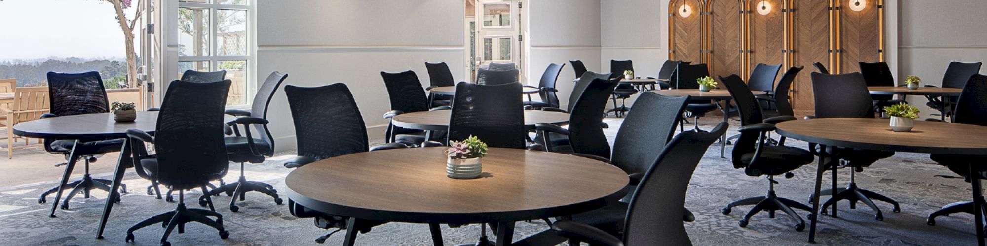 The image shows a modern meeting room with round tables, black chairs, large circular chandeliers, and wood beam ceiling accents.