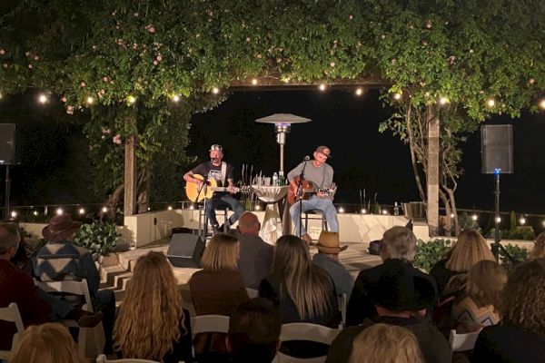 Two musicians are performing on a small outdoor stage surrounded by greenery, with an audience seated and watching.