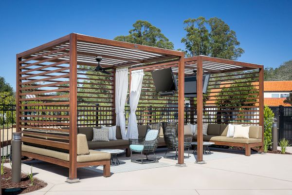 A modern outdoor seating area with two shaded pergolas featuring cushioned seats, a mounted TV, and small tables on a sunny day.