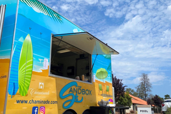 A vibrant food truck with beach-themed decor and social media icons is parked outdoors, near a pool sign and buildings, under a partly cloudy sky.