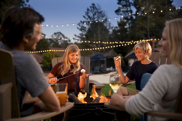 A group of four people sits around a fire pit, enjoying roasted marshmallows and drinks, with string lights overhead and trees in the background.