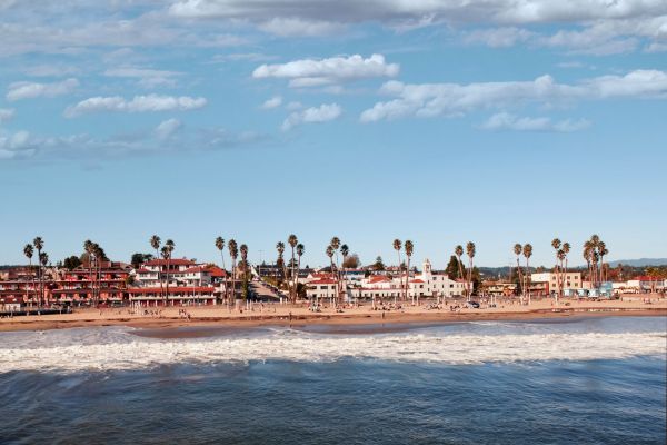 A coastal scene with waves hitting the sandy shore, lined with palm trees and buildings in the background under a cloudy sky.