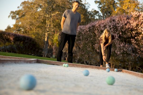 Two people are playing bocce ball on an outdoor court surrounded by trees and bushes in a sunny setting.