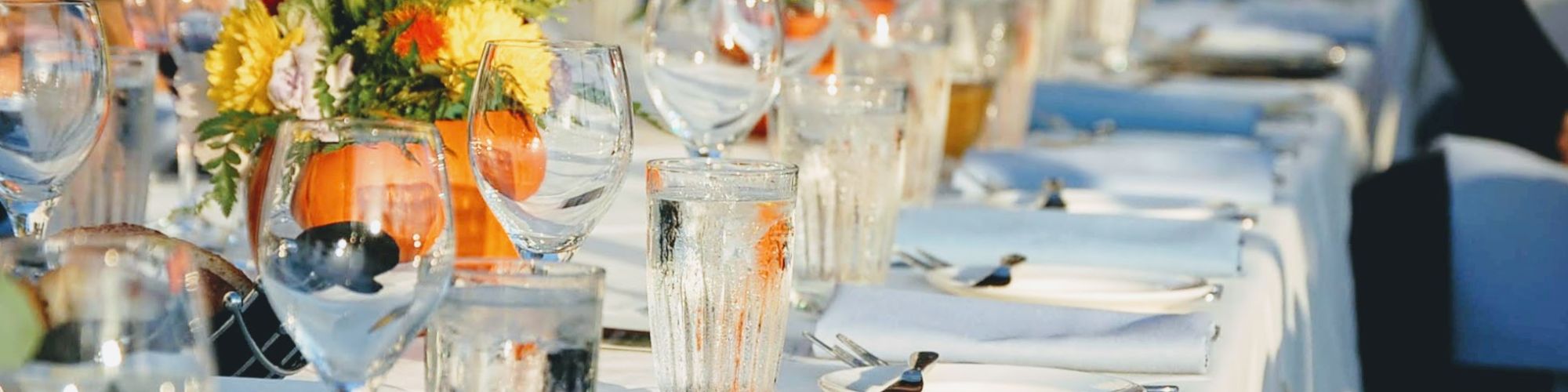 A long table is set for a formal event with white tablecloths, glassware, cutlery, and flower centerpieces placed decoratively.