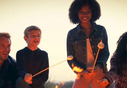 A group of four people are gathered around a campfire, smiling and roasting marshmallows on sticks.