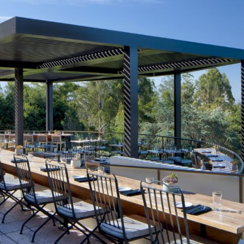 An outdoor dining area with tables and chairs is set under a large pergola, surrounded by trees and greenery, offering scenic views.