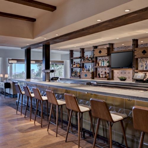 This image shows an empty bar with stylish chairs, a tiled backsplash, a large countertop, a TV, and shelves stocked with bottles, set in a cozy interior.