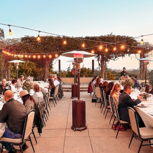 The image shows a group of people dining at an outdoor restaurant under string lights and heaters, with trees and a sunset in the background.