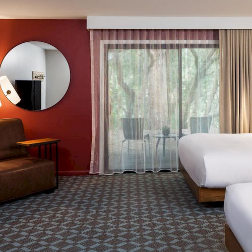 A hotel room with two beds, a brown sofa, a round mirror, a floor lamp, and a balcony with a view of trees outside, under a red accent wall.