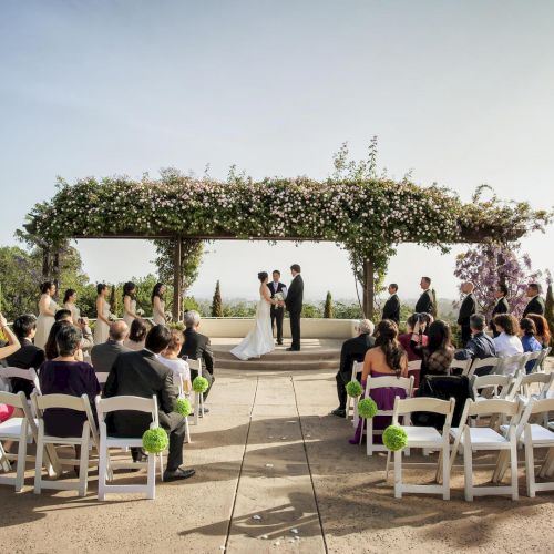 A wedding ceremony is taking place outdoors with guests seated, a couple standing under a floral arch, and a clear sky in the background.