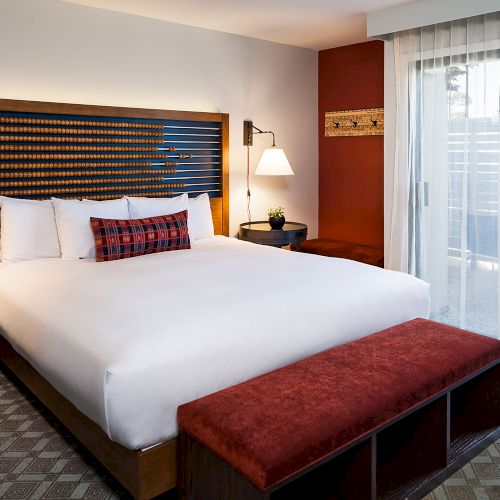 A neatly arranged hotel room features a large bed with white linens, a red bench at the foot, a side table with a red lamp, and a window with sheer curtains.