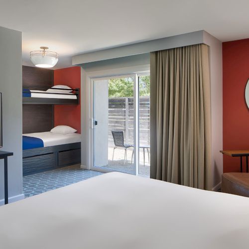 The image shows a modern hotel room with a bunk bed, a large bed, a TV, a sliding glass door, a brown chair, and a round mirror on a red wall.