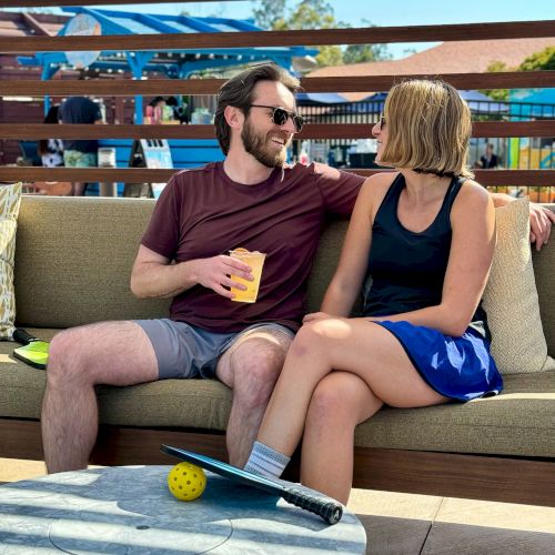 Two people are sitting on a couch outdoors, smiling at each other. A pickleball paddle and ball are on the table in front of them, with a lively background.