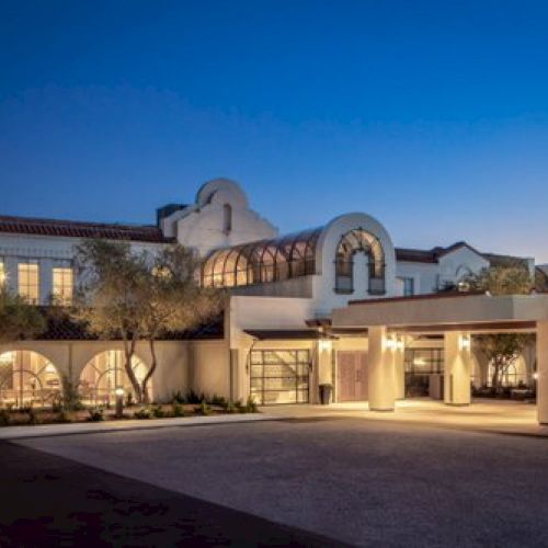 The image shows a well-lit building at twilight with Mediterranean architectural elements, including arched windows and clay tile roofs.