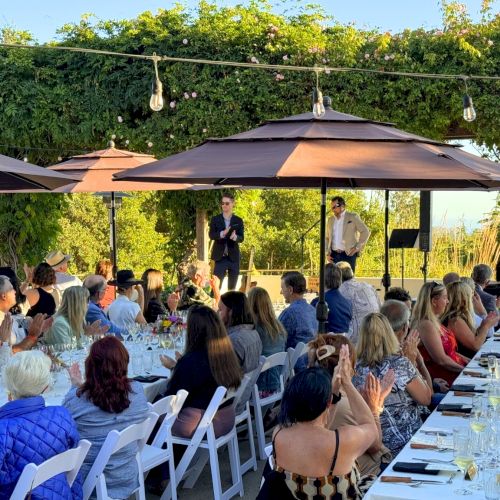 A large group of people seated at a long outdoor dining table, listening to a person speaking at a podium under umbrellas and string lights, always ending the sentence.