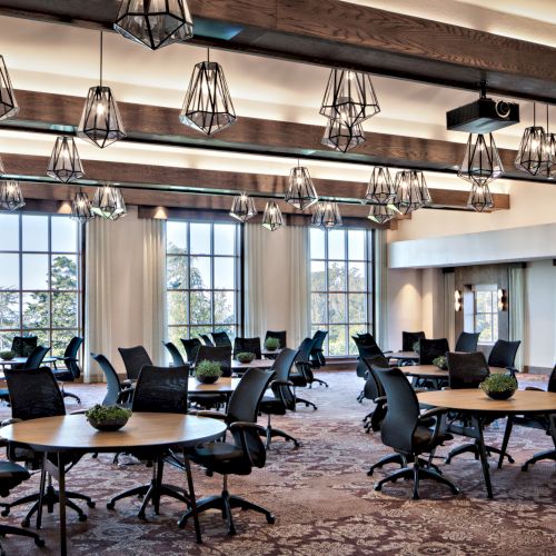 A modern conference room with round tables, ergonomic chairs, decorative lights on a wooden-beamed ceiling, large windows, and potted plants on tables.