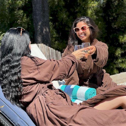 Two people in brown robes are toasting glasses while lounging outdoors on a sunny day, surrounded by greenery and trees, and appearing to enjoy.