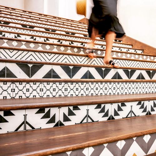 The image shows a person in a black outfit walking up a staircase decorated with black and white geometric patterned tiles.