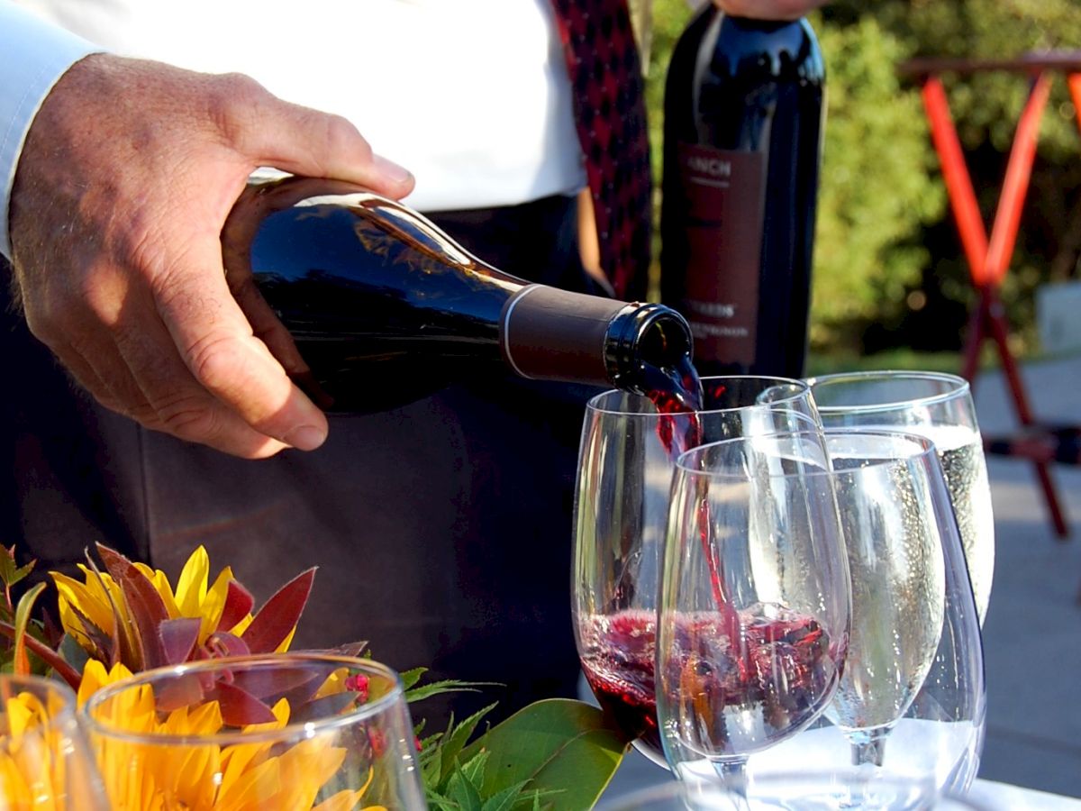 A person is pouring red wine into glasses at an outdoor setting with flowers and a background of blurred greenery.