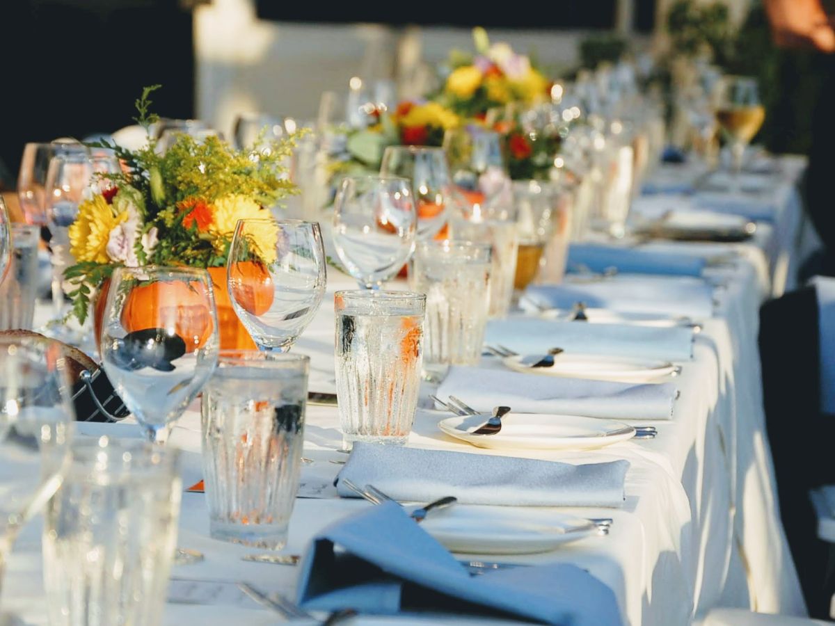 A long, elegantly set dining table with white linens, floral centerpieces, and glassware, ready for a formal or celebratory event.