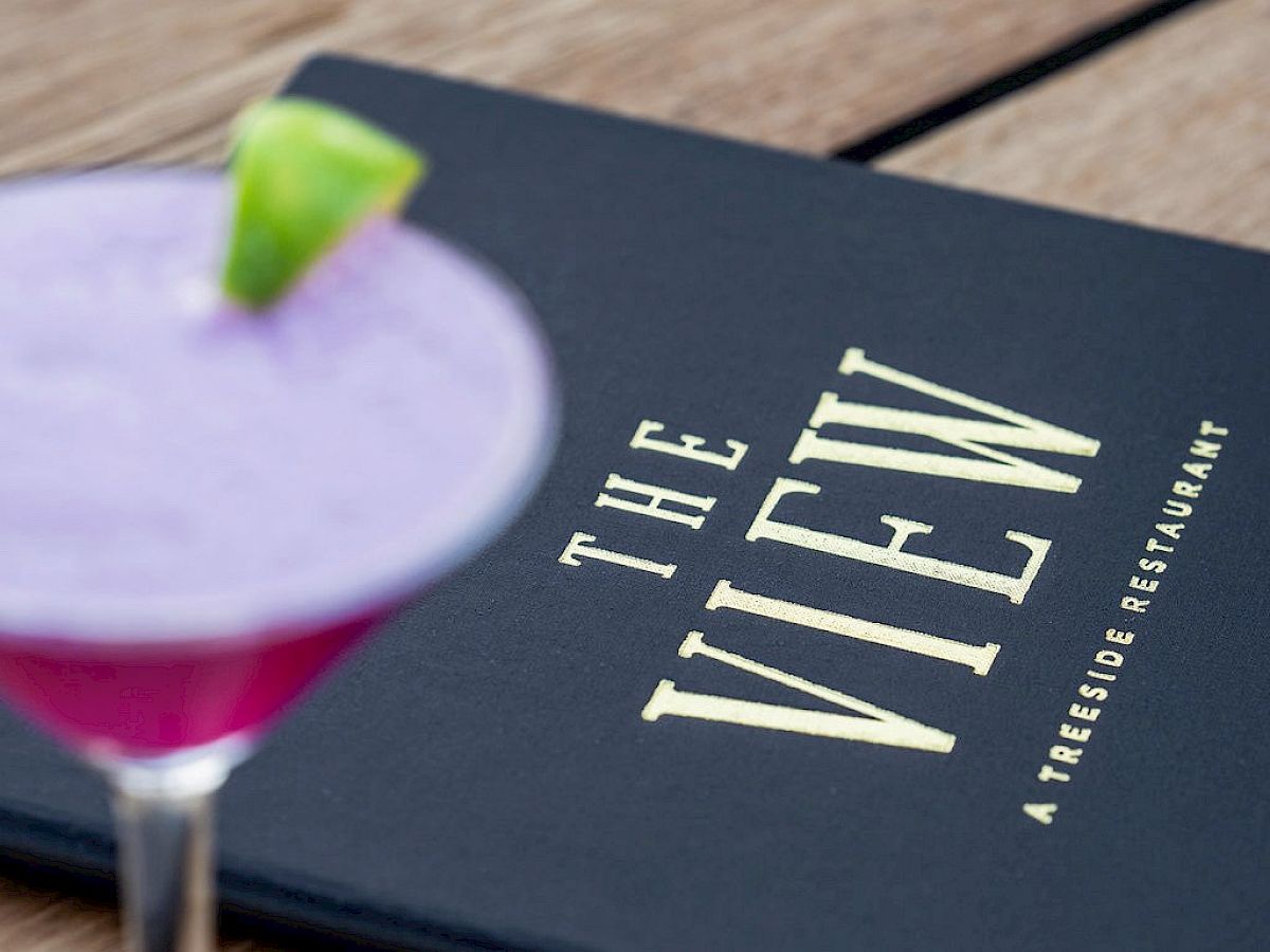 A purple cocktail with a lime wedge sits on a wooden table next to a dark menu titled 