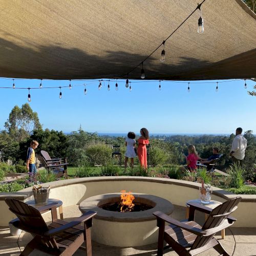 A group of people enjoys an outdoor patio with a fire pit, shaded by a canopy and decorated with hanging lights, overlooking a scenic, green landscape.