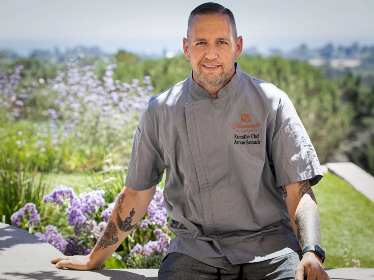 A person wearing a chef's jacket, seated outdoors with a scenic, floral background, and has tattoos on their arms.