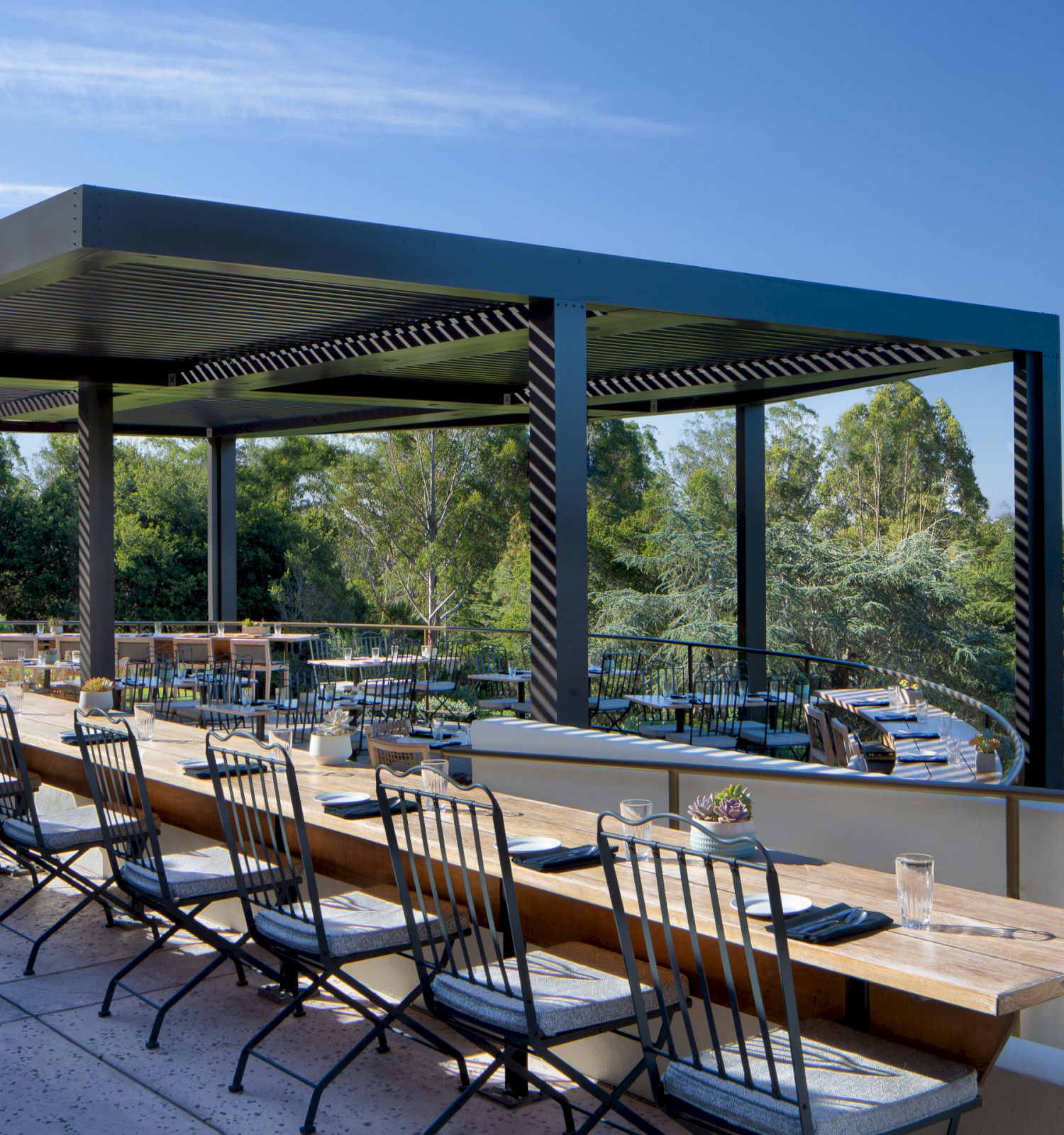 An outdoor terrace with a pergola, featuring a long wooden dining table and chairs, overlooks lush greenery and trees against a clear blue sky.