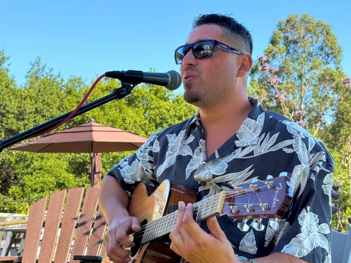 A person wearing sunglasses and a floral shirt plays an acoustic guitar and sings into a microphone outdoors, with trees in the background.