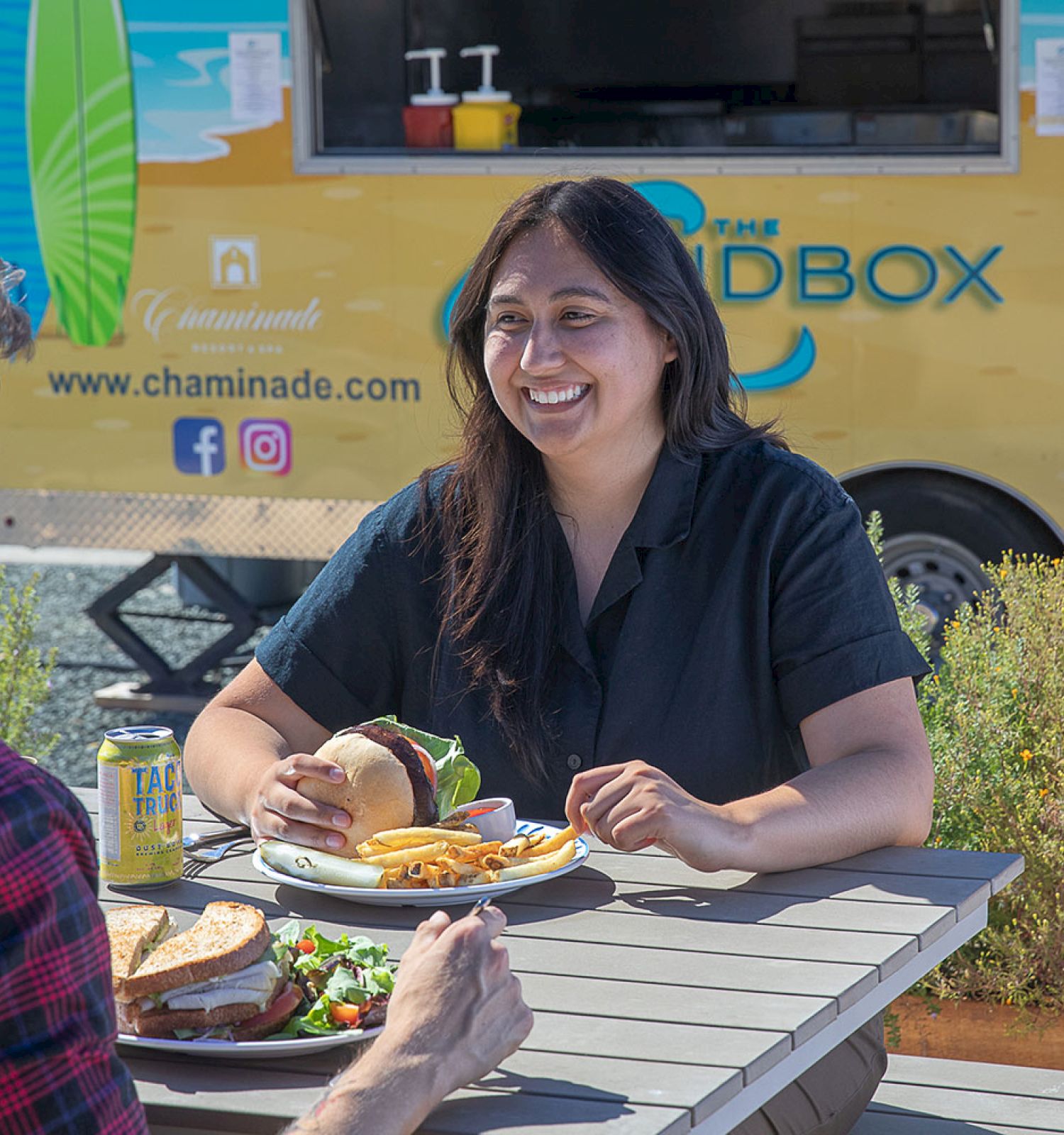 Two people are enjoying a meal together at an outdoor table near a food truck on a sunny day, with one person smiling and eating a sandwich.