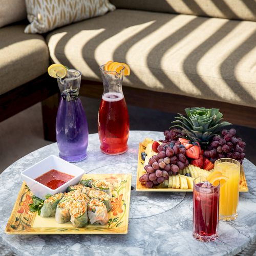 A marble table with spring rolls and sauce, fruit platters, and colorful drinks in a bright outdoor setting.