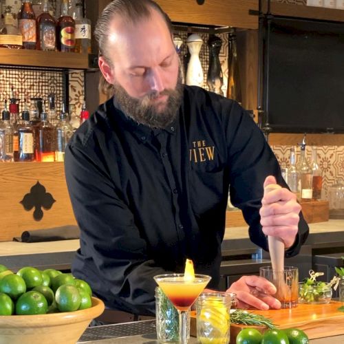 A bartender is preparing a drink at a bar with a bowl of limes beside him and various bottles in the background, ending the sentence.