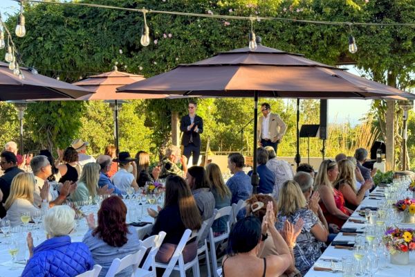 A group of people is attending an outdoor event or celebration with dining tables, under umbrellas, as a speaker addresses the audience.